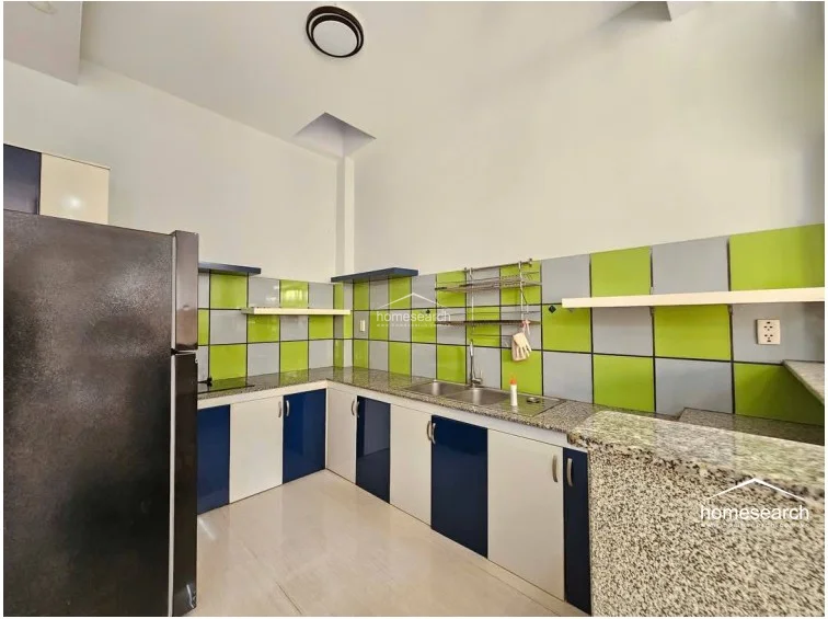 The kitchen features a functional U-shaped layout with a granite countertop and a distinctive checkerboard tile backsplash in green and grey. It is equipped with a large refrigerator, stainless steel sink, and ample under-counter cabinet storage, maintaining a clean and organized appearance.