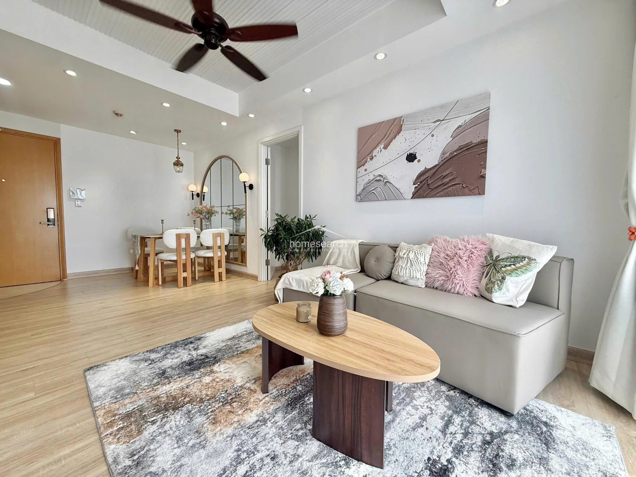 This open-concept living and dining area features a cohesive, neutral color palette with light wood-look flooring and crisp white walls. The space is anchored by a modern ceiling fan, recessed lighting, and a striking arched floor-to-ceiling mirror that enhances the sense of depth in the dining zone.