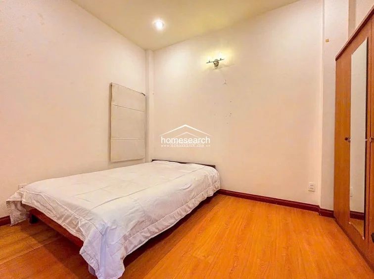 This bedroom features warm wood-look laminate flooring and a minimalist layout centered around a double bed. The room is finished with neutral cream walls, a single wall-mounted light fixture, and a standalone wooden wardrobe, presenting a clean and well-maintained living space.