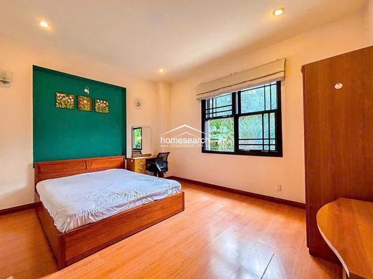 This spacious bedroom is highlighted by a vibrant green accent wall behind the bed, which contrasts with the neutral walls and wood-look flooring. It includes a functional wooden wardrobe, a vanity desk with a chair, and a large window with a roman shade that offers a view of lush greenery, providing good natural light.