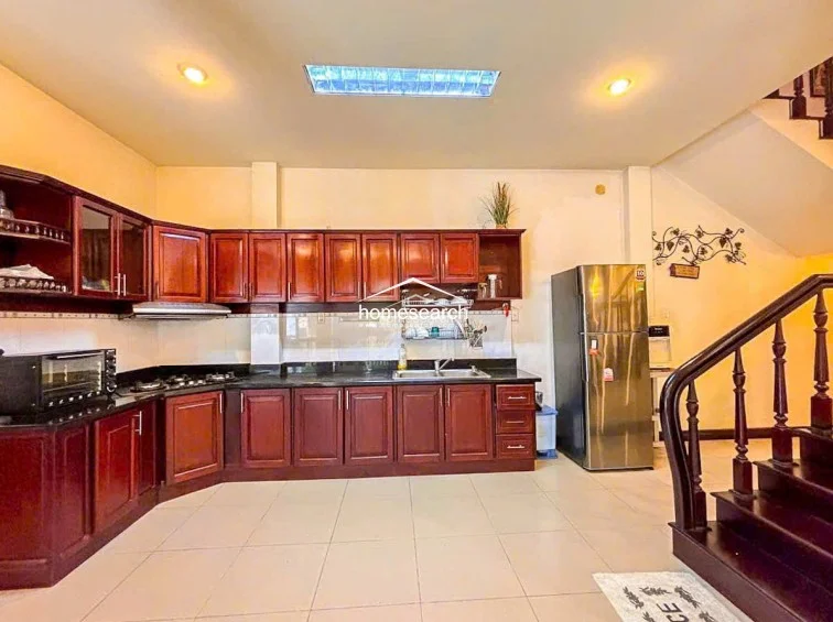 This kitchen workspace highlights a functional U-shaped layout with ample dark wood cabinetry and a large stainless steel refrigerator. The space is well-lit by recessed lighting and a central skylight, and it connects directly to a wooden staircase, maintaining a consistent aesthetic with the rest of the home.
