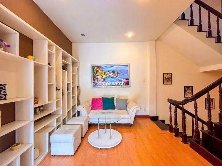 This living area serves as a central hub, featuring an open-plan design that connects directly to an upper floor via a dark wood staircase. The space is highlighted by a large, floor-to-ceiling built-in shelving unit, a neutral-toned sofa, and light wood flooring that creates a bright, welcoming atmosphere.