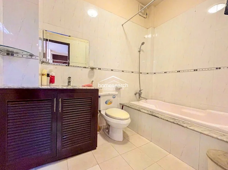 This bathroom is equipped with a standard bathtub-shower combination and a dark wood vanity with louvered cabinet doors. The space is fully tiled in a light, neutral color scheme, appearing functional and well-maintained with basic fixtures.