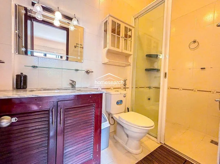 This bathroom features a modern glass-enclosed shower stall and a vanity unit with a marble countertop and dark wood cabinetry. The room is brightened by a decorative vanity mirror with integrated lighting and wall-mounted storage shelves, maintaining a clean and organized appearance.