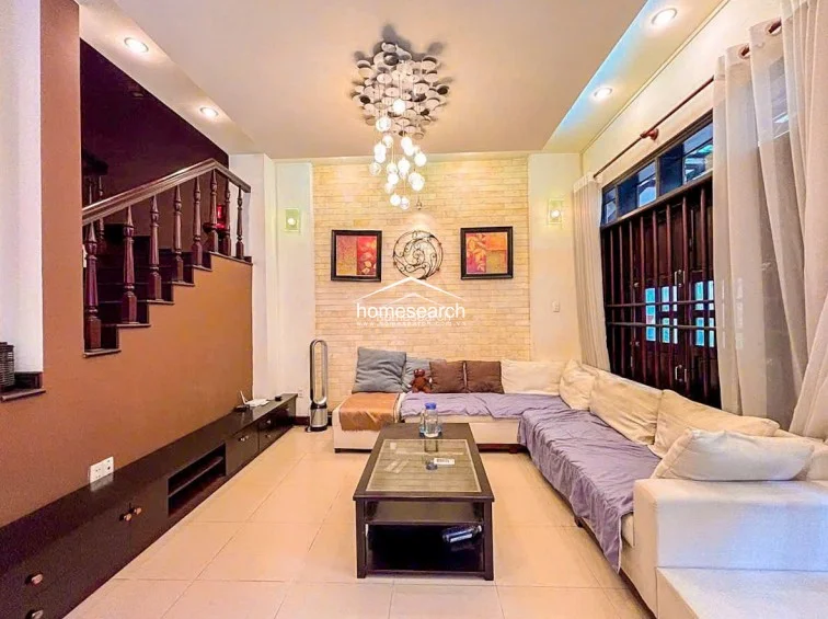 The living room serves as a comfortable gathering space, anchored by a large L-shaped sofa and a modern coffee table set against a decorative stone-textured feature wall. A striking, multi-bulb chandelier provides a focal point, while the dark wood staircase and matching low-profile media console add architectural depth to the room.