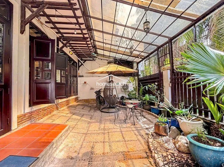 This outdoor terrace features a covered patio with a translucent roof, providing a sheltered transition space between the interior and the garden. The area is paved with natural stone tiles and includes a cozy seating arrangement with an umbrella, hanging chair, and lush potted plants, creating a serene, well-maintained relaxation spot.