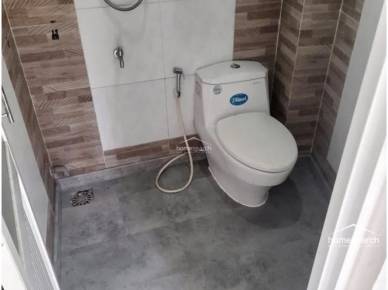 This compact bathroom features a white pedestal sink, a wall-mounted mirror with a glass shelf, and a separate enclosed toilet area. The walls are finished with white and brown decorative tiles, and the floor is covered in dark grey non-slip tiles.