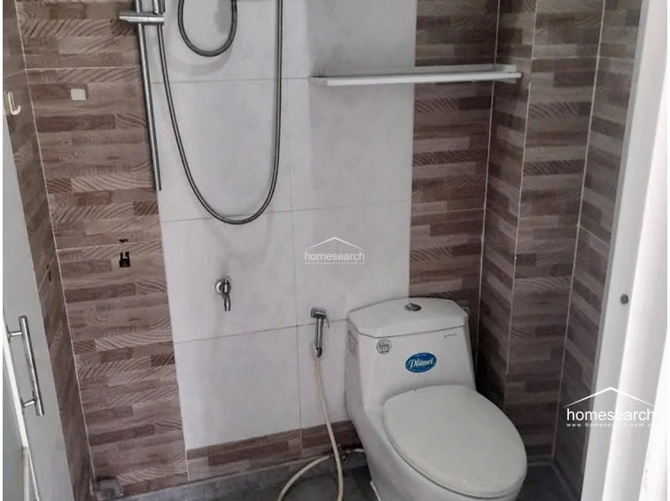 This bathroom features a compact, functional layout with a white porcelain toilet and a handheld shower fixture. The walls are finished with a combination of plain white tiles and wood-look accent tiles, and a simple shelf is mounted for storage.