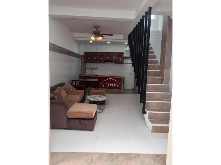 This combined living and kitchen area utilizes an open floor plan with white tiled flooring and a prominent staircase featuring brown, wood-patterned tile steps. The kitchen is equipped with dark wood cabinetry, and the room is cooled by a ceiling fan, maintaining a cohesive, functional flow throughout the ground level.