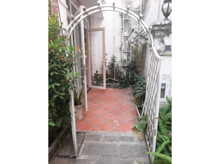 This image captures a private outdoor terrace or entrance area featuring terracotta-style floor tiles and a decorative metal archway. The space is enclosed by lush plants and greenery, creating a secluded, garden-like transition point that connects to the interior through a glass-paneled door.