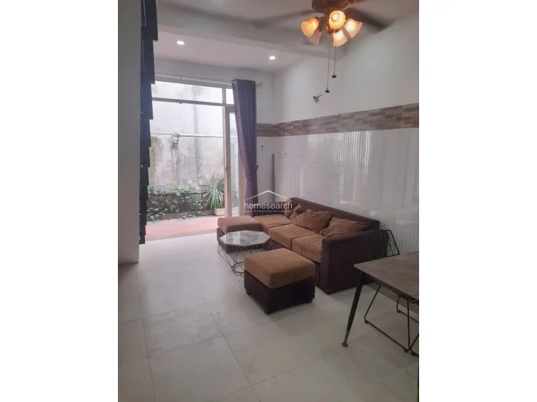 This living area features a brown sofa set and a small dining table, with light-colored tile flooring throughout. A large glass door provides natural light and leads to an outdoor patio area with greenery, while a ceiling fan with light fixtures is mounted above.