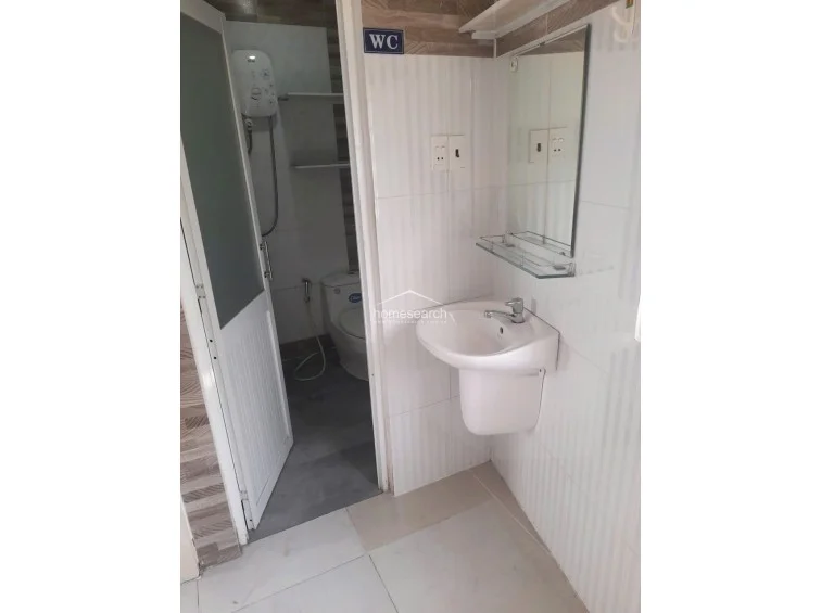 This bathroom layout includes a wall-mounted sink and mirror, with a separate door leading to the toilet and shower area. The space is finished with light-colored wall tiles and a dark grey tiled floor, and it includes a wall-mounted water heater for the shower.