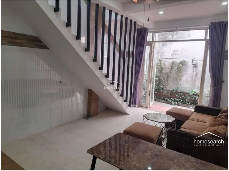This open-concept living area features a modern staircase with a striking black-and-white vertical slat railing that serves as a visual centerpiece. The space is brightened by a large floor-to-ceiling glass door that offers a view of the outdoor greenery, while the neutral-toned sofa and marble-topped coffee table provide a comfortable seating arrangement.
