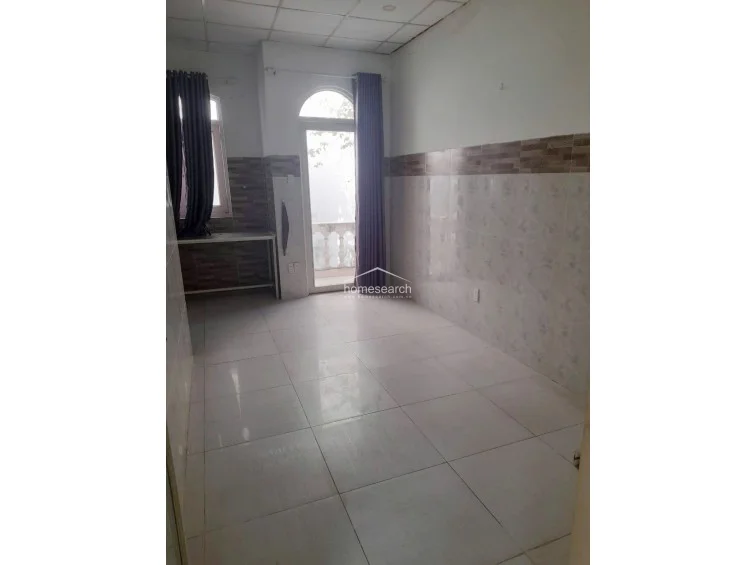 This room appears to be a multi-purpose space or bedroom featuring white tiled walls with a decorative brown accent border and matching floor tiles. It includes a glass-paned door that opens to a small balcony, allowing for natural light, and a simple metal table frame is positioned against the wall.