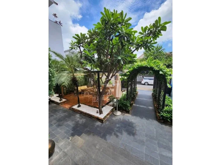 This outdoor courtyard features a paved stone patio with a central seating area shaded by a mature tree and a large umbrella. The space is framed by a lush, vine-covered metal archway that provides a charming transition to the property’s entrance.