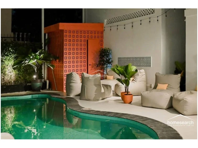 This poolside lounge area offers a relaxed, resort-style atmosphere with comfortable beanbag seating and ambient string lighting. The space includes a distinctive orange breeze-block structure and is surrounded by well-maintained tropical greenery, creating a private oasis.