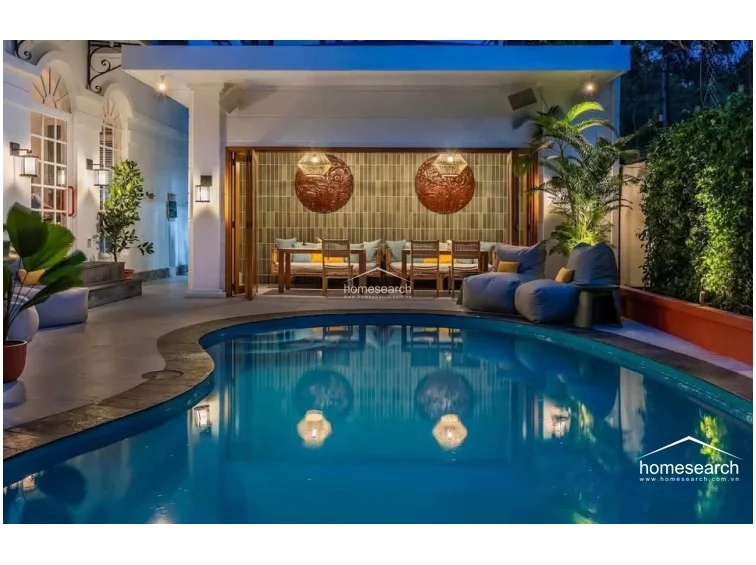 This outdoor lounge area features a kidney-shaped swimming pool and a covered terrace that seamlessly connects to the interior through folding glass doors. The space is furnished with comfortable beanbag chairs and a dining set, complemented by warm wall lighting and lush greenery for a private, resort-like atmosphere.