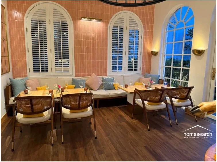 This dining space features a sophisticated blend of warm wood flooring and soft pink vertical wall tiles, complemented by elegant arched windows with plantation shutters. The layout includes built-in banquette seating and mid-century modern style cane-back chairs, creating a bright, inviting flow filled with natural light.