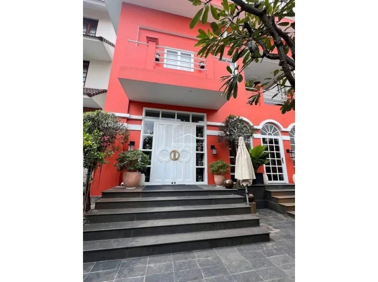This striking exterior view showcases the property’s bold coral facade, contrasted by white trim and elegant arched windows. The grand entrance is elevated by a wide stone staircase, framed by potted plants and a balcony above, suggesting a well-maintained and architecturally unique residence.