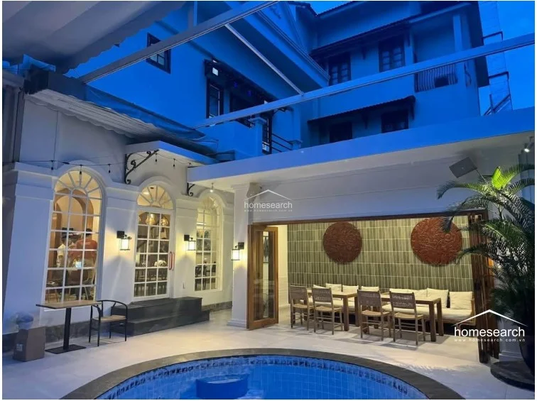 This evening view highlights the property's seamless indoor-outdoor flow, with the illuminated terrace dining area opening directly onto the pool deck. The architecture features classic white columns and arched windows, creating a serene and well-maintained environment perfect for evening relaxation.