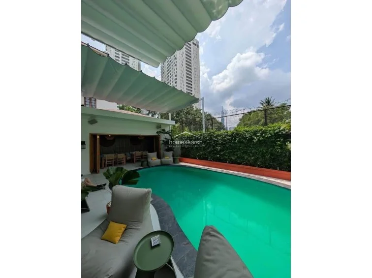 This outdoor space is defined by a large, kidney-shaped pool and a retractable awning that provides flexible shade for the lounge area. The perimeter is enclosed by a high, well-maintained hedge, ensuring privacy while offering a view of the surrounding urban skyline.