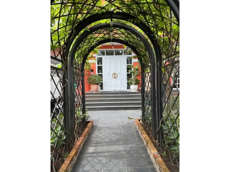 The main entrance is highlighted by a grand, white double-door design with circular gold hardware, set against a vibrant coral-colored exterior wall. A stone-paved path leads through a decorative metal archway, climbing a short set of stairs to the elevated entryway.