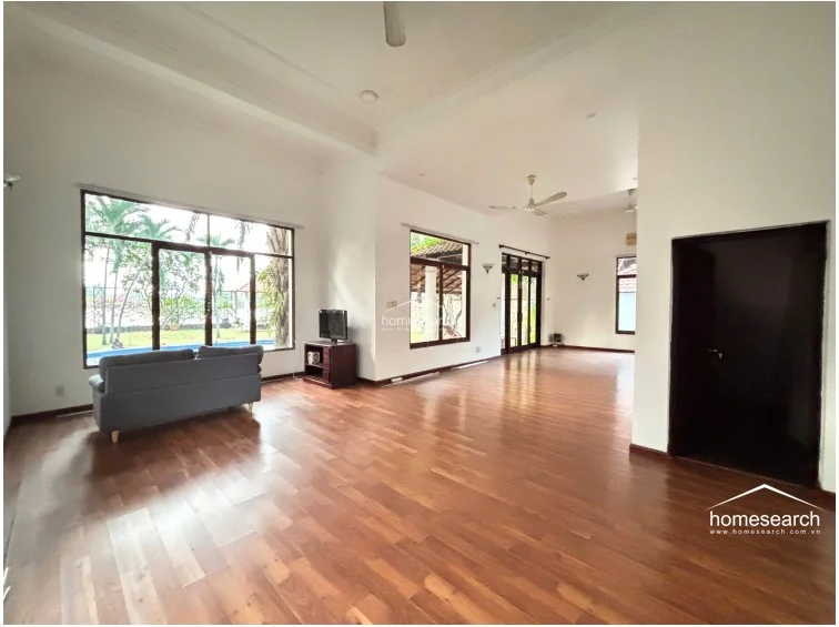 This perspective highlights the expansive layout of the main living area, which flows seamlessly toward a large window overlooking a private garden and pool area. The space is equipped with ceiling fans and a wall-mounted AC unit, maintaining a bright and airy atmosphere throughout the neutral-toned interior.