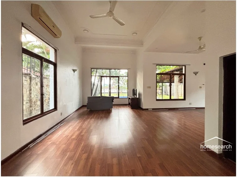 This living room area showcases the home's open flow, connecting the main lounge to the rest of the residence through wide, unobstructed passages. The space is well-lit by large windows, features a wall-mounted AC unit, and is finished with polished wood flooring and crisp white walls, reflecting a well-maintained condition.