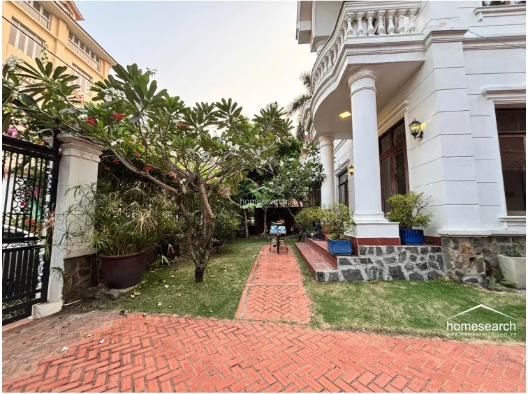 This image displays the front entrance area of a neoclassical-style villa, featuring a prominent white facade with classical columns and a balustraded balcony above. The exterior is well-maintained, with a lush garden, mature trees, and a terracotta-tiled pathway leading to the main entrance.