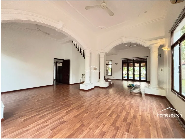 This expansive open-concept living area is defined by grand architectural elements, including classical columns and elegant archways that create a seamless flow between spaces. The room features warm wood flooring, high ceilings with decorative molding, and large windows that provide abundant natural light and views of the outdoor pool area.