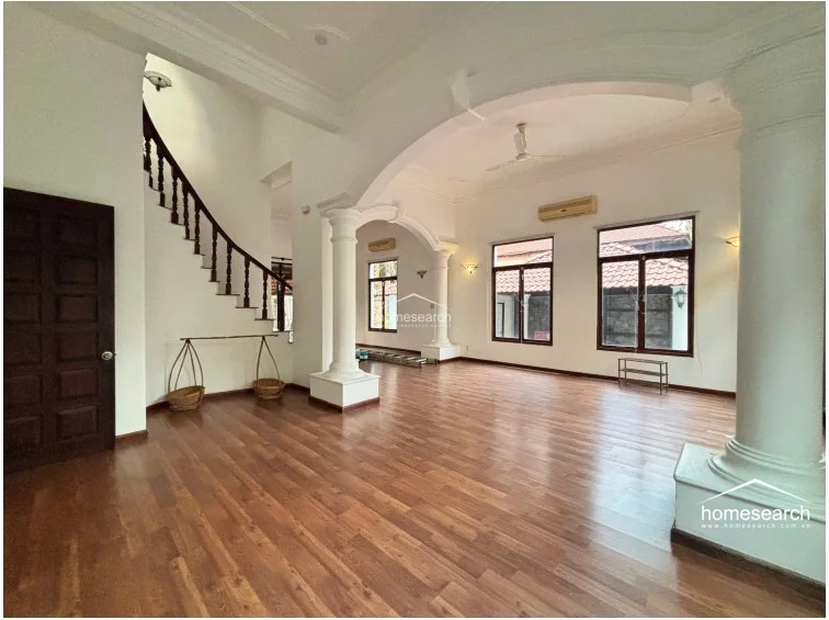 This grand living space showcases a sophisticated design with a sweeping staircase featuring a dark wood banister and white spindle railings. The room is characterized by its airy, open layout, white walls, and large windows, all complemented by warm-toned wooden flooring and wall-mounted air conditioning units for climate control.