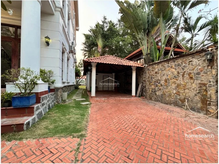 This side view highlights the property's gated driveway and a covered carport with a traditional terracotta-tiled roof. The area is enclosed by a rustic stone wall and surrounded by dense tropical landscaping, indicating a private and well-secured outdoor space.