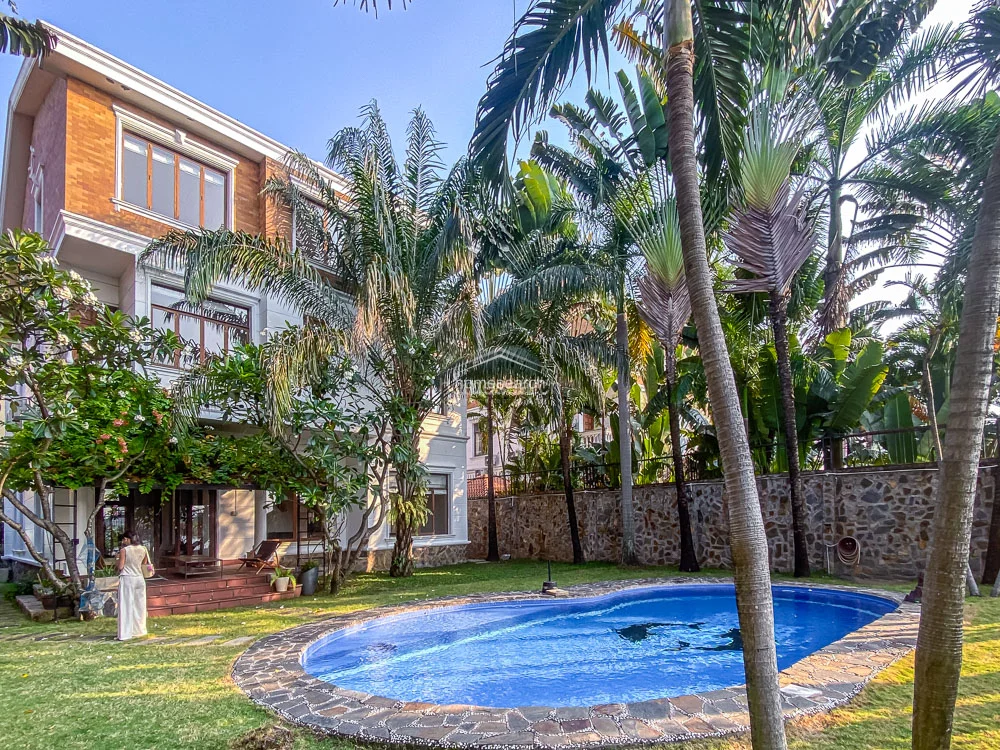 This image showcases a multi-story villa featuring a classic architectural style with warm-toned brick accents and large, grid-paned windows that invite ample natural light. The exterior flows seamlessly into a lush, tropical garden setting, highlighted by a kidney-shaped swimming pool and mature palm trees. The property appears well-maintained, with stone-clad boundary walls and a spacious terrace area providing a serene outdoor living environment.