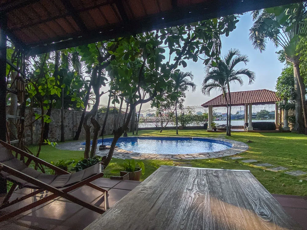 This view highlights the tranquil outdoor living space, featuring a covered patio area with a wooden deck that overlooks the private pool and garden. The open-air layout provides a direct connection between the indoor living space and the recreational area, offering a perfect spot for relaxation under the shade of mature trees. The property maintains a high level of privacy with a stone perimeter wall and a charming gazebo structure in the distance.