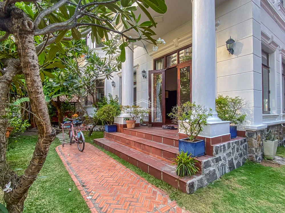 This perspective captures the grand entrance of the villa, characterized by imposing white columns and a raised porch finished with terracotta-style tiling. The layout includes a direct transition from the garden path to the main living area through double glass doors, framed by vibrant potted plants. The exterior walls feature a clean, light-colored finish that contrasts elegantly with the rustic stone base and the natural greenery of the surrounding landscape.