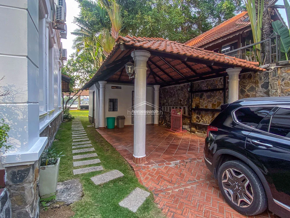 This image displays a functional carport area with a traditional tiled roof and sturdy, white-painted columns that mirror the villa's architectural aesthetic. The space is paved with durable terracotta tiles, providing a seamless flow from the driveway to the side garden path. The area is well-integrated into the property's design, offering convenient vehicle storage while maintaining the cohesive, tropical-inspired look of the home.