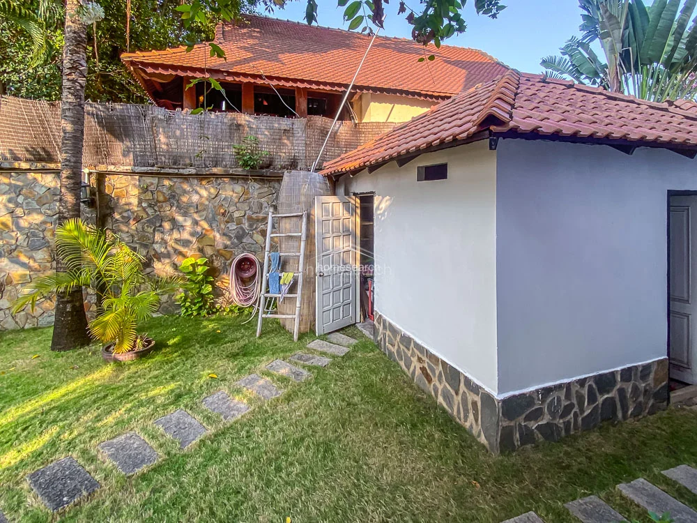 This shot focuses on a secondary utility or storage building located within the garden, featuring a simple, clean design with a white exterior and stone foundation. The structure is surrounded by a well-manicured lawn and stone stepping stones, ensuring easy access from the main residence. The overall condition of the outdoor structures appears excellent, reflecting a well-cared-for property with thoughtful landscaping and functional auxiliary spaces.