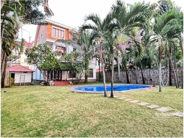This exterior view showcases a multi-story residence featuring a classic brick facade and a spacious, lush garden. The property includes a private, kidney-shaped swimming pool and a detached outbuilding, all surrounded by mature palm trees and a stone perimeter wall.