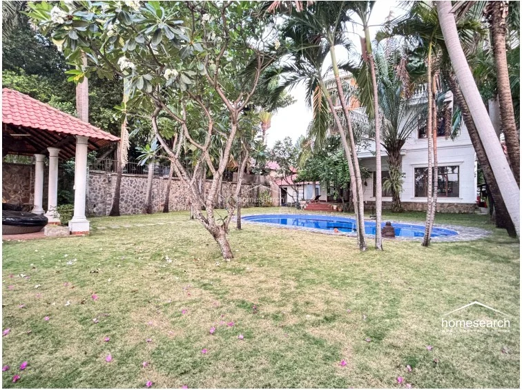 This angle highlights the expansive lawn and the serene pool area, framed by vibrant tropical foliage and palm trees. A charming, open-air pavilion with a terracotta-tiled roof provides a perfect shaded spot for outdoor lounging or entertaining.