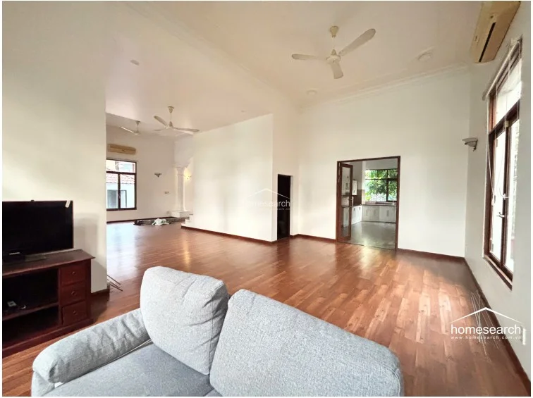 This living room space is bright and airy, featuring high ceilings, ceiling fans, and wall-mounted air conditioning units for climate control. The layout flows effortlessly into a separate kitchen area, while large windows ensure the space remains well-lit and connected to the outdoor environment.