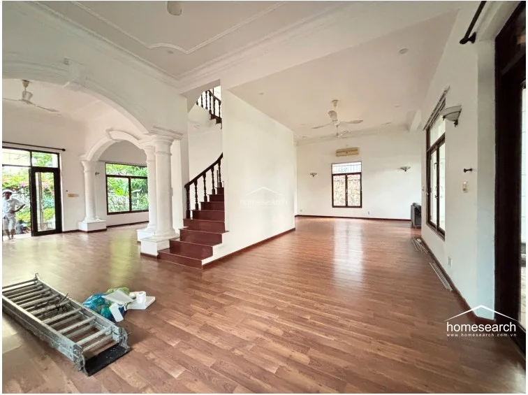 This spacious open-plan living area features high ceilings with ornate crown molding and elegant architectural columns that define the transition to the staircase. The room is finished with warm-toned wood-look flooring, white walls, and multiple large windows that provide ample natural light and views of the surrounding greenery.