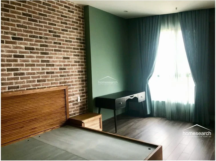 This bedroom creates a cozy, textured atmosphere with an exposed brick feature wall and deep green painted walls. It is furnished with a slatted wood headboard, a minimalist black desk, and dark wood flooring, all illuminated by soft natural light filtering through floor-length curtains.