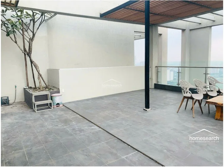 This outdoor terrace provides a generous, open-air space finished with large-format grey tiles and partially shaded by a slatted wood pergola. It features a built-in planter with a mature tree, glass railings for unobstructed city views, and a simple patio furniture set, making it an ideal area for relaxation.