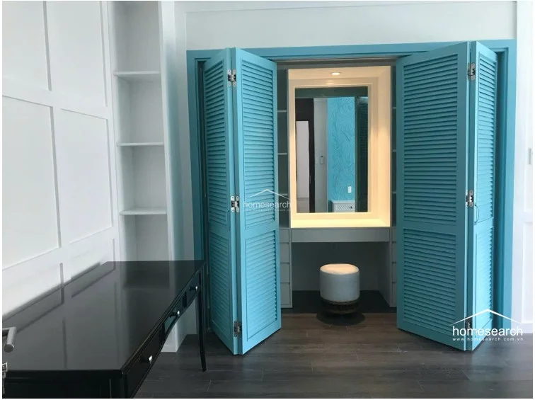 This dedicated dressing area or walk-in closet is highlighted by a vibrant turquoise louvered cabinet that opens to reveal a vanity station with a mirror and stool. The space is complemented by white paneled walls and a sleek black desk, offering a clean, organized aesthetic with ample storage.