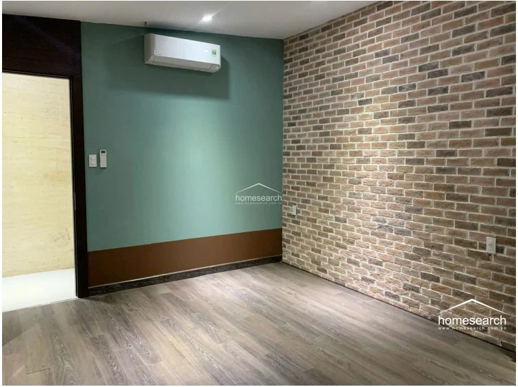 This bedroom space showcases a trendy industrial-inspired accent wall with textured brick veneer paired with a bold sage green wall. It includes light wood-look flooring, a wall-mounted AC unit, and a doorway leading to an adjacent room, suggesting a functional and contemporary layout.