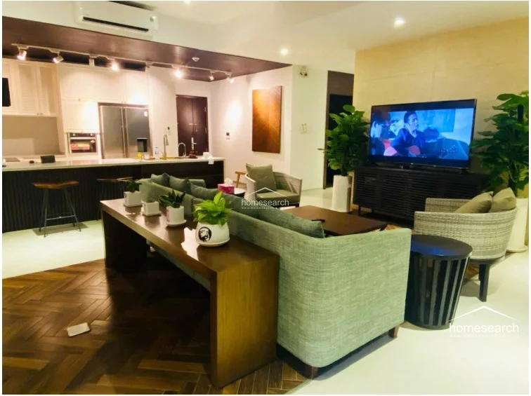 This open-concept living and kitchen area features a sophisticated blend of dark wood herringbone flooring and sleek white surfaces. The kitchen is equipped with modern built-in appliances and a breakfast bar, while the lounge is furnished with contemporary seating and a large wall-mounted television.