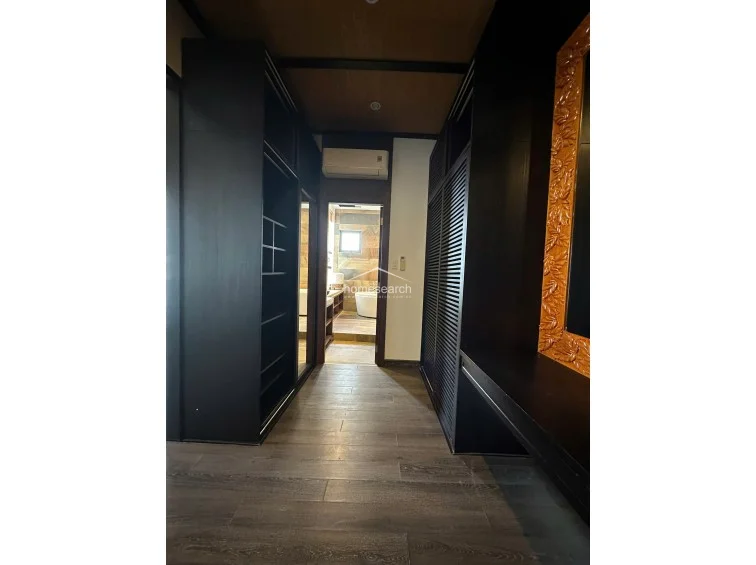 This transition hallway serves as a walk-in wardrobe, featuring floor-to-ceiling dark wood cabinetry with louvered doors and open shelving for maximum storage. The space leads directly into a bathroom visible at the end, and the dark wood theme is carried through the flooring and ceiling, creating a cohesive, moody atmosphere.