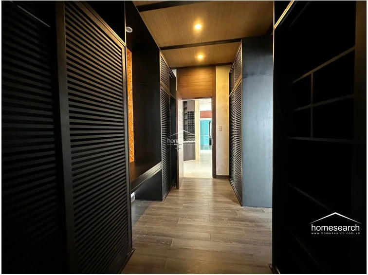 This walk-in closet or dressing area is defined by dark, louvered cabinetry that provides ample storage space. The room features warm wood-paneled ceilings and flows directly into an adjacent living space, maintaining a cohesive and modern aesthetic.