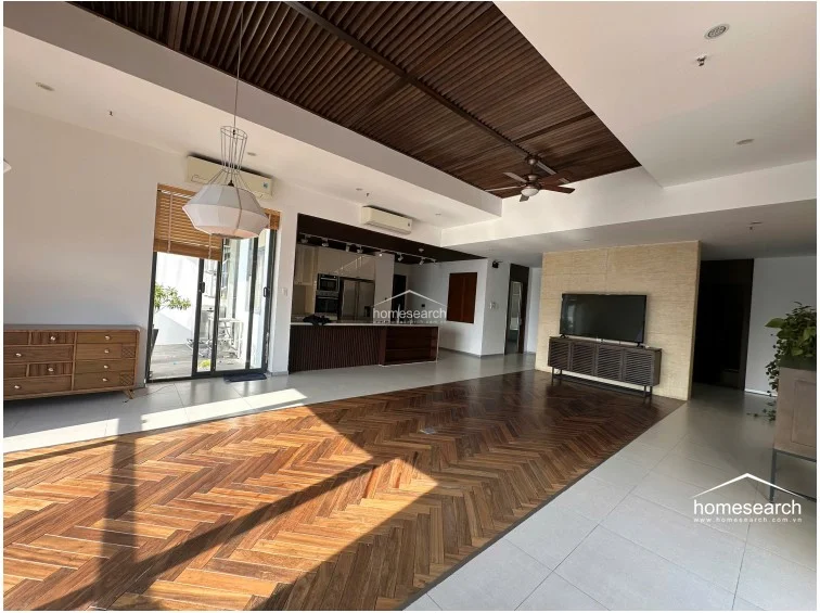 This expansive open-plan living area features a sophisticated combination of herringbone-patterned wood flooring and light-colored tile. The space includes a high-end kitchen with a breakfast bar, a ceiling fan, and a wall-mounted AC unit, all illuminated by a modern pendant light and recessed lighting.