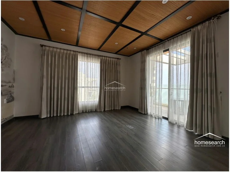 This spacious room features a striking coffered ceiling with wood-slatted panels and dark framing, adding architectural depth. Large floor-to-ceiling windows with neutral curtains provide ample natural light and access to a balcony, while dark wood-style flooring grounds the space.